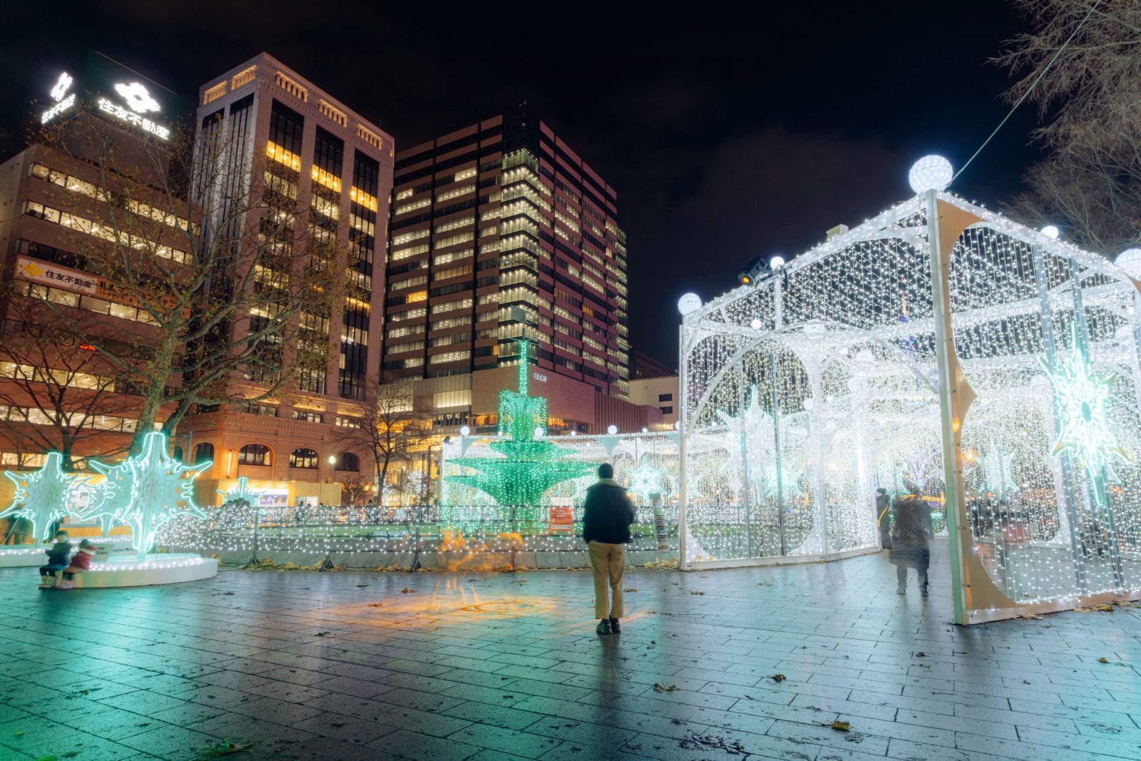 A white illuminated passage beside green light sculptures and city buildings.
